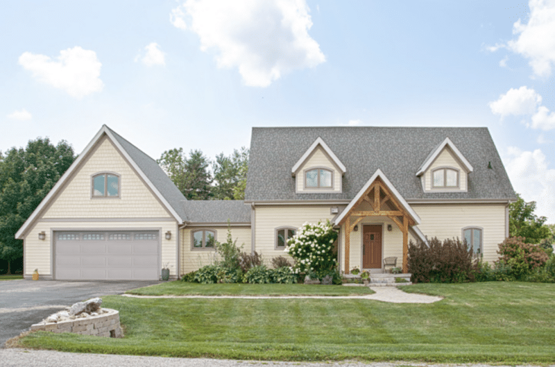 A charming, cream-colored house featuring a central A-frame entrance with wooden details and two flanking side sections, each with dormer windows. The house has a large attached garage with a white door, and is surrounded by a well-maintained lawn with a gravel path leading to the ornate front door, set against a backdrop of lush greenery and a clear sky